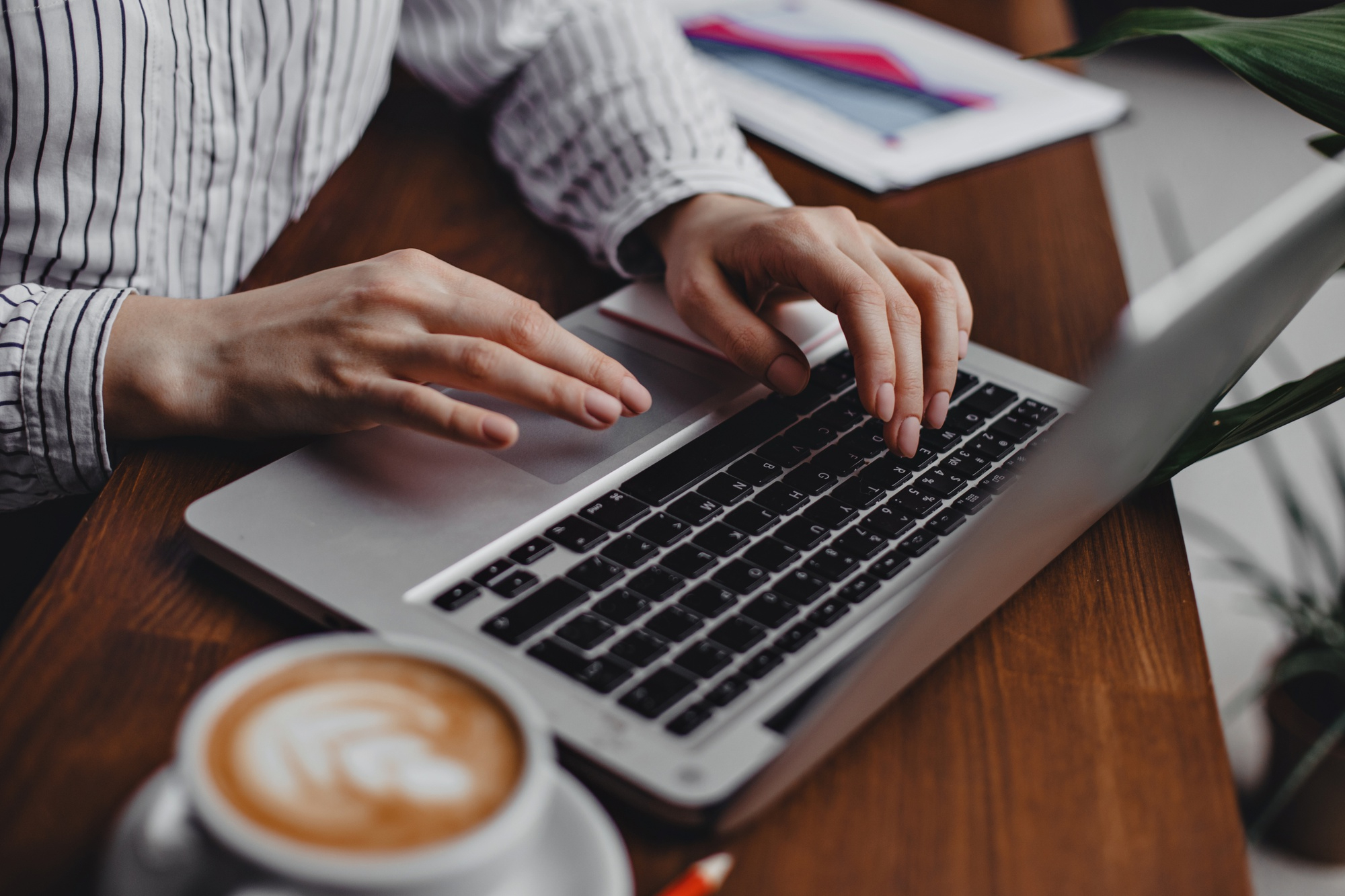 female-hands-press-laptop-keys-while-sitting-wooden-table-with-white-cappuccino-cup