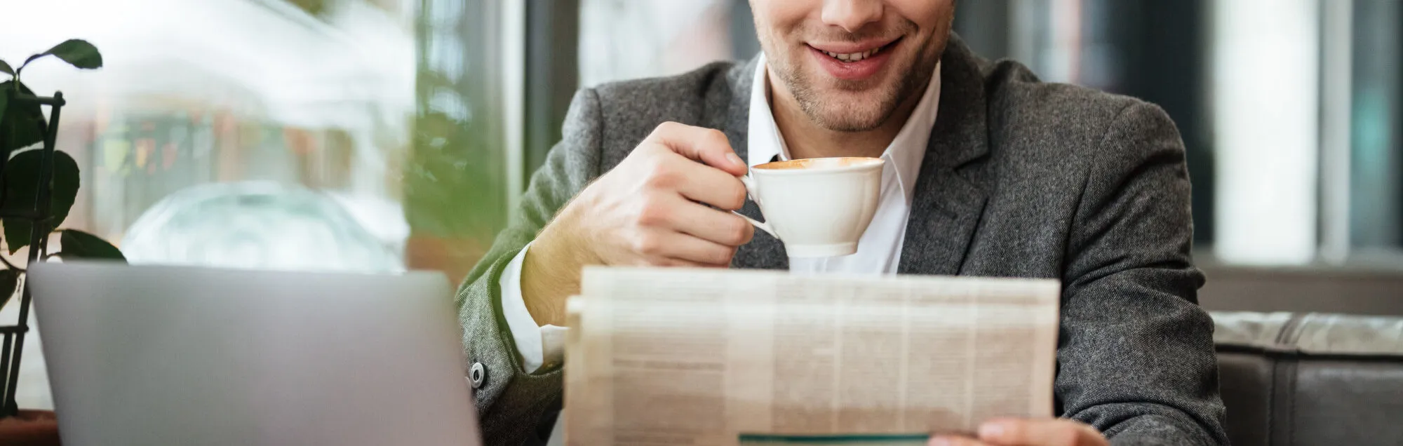 smiling-businessman-sitting-by-table-cafe-with-laptop-computer-while-reading-newspaper-drinking-coffee